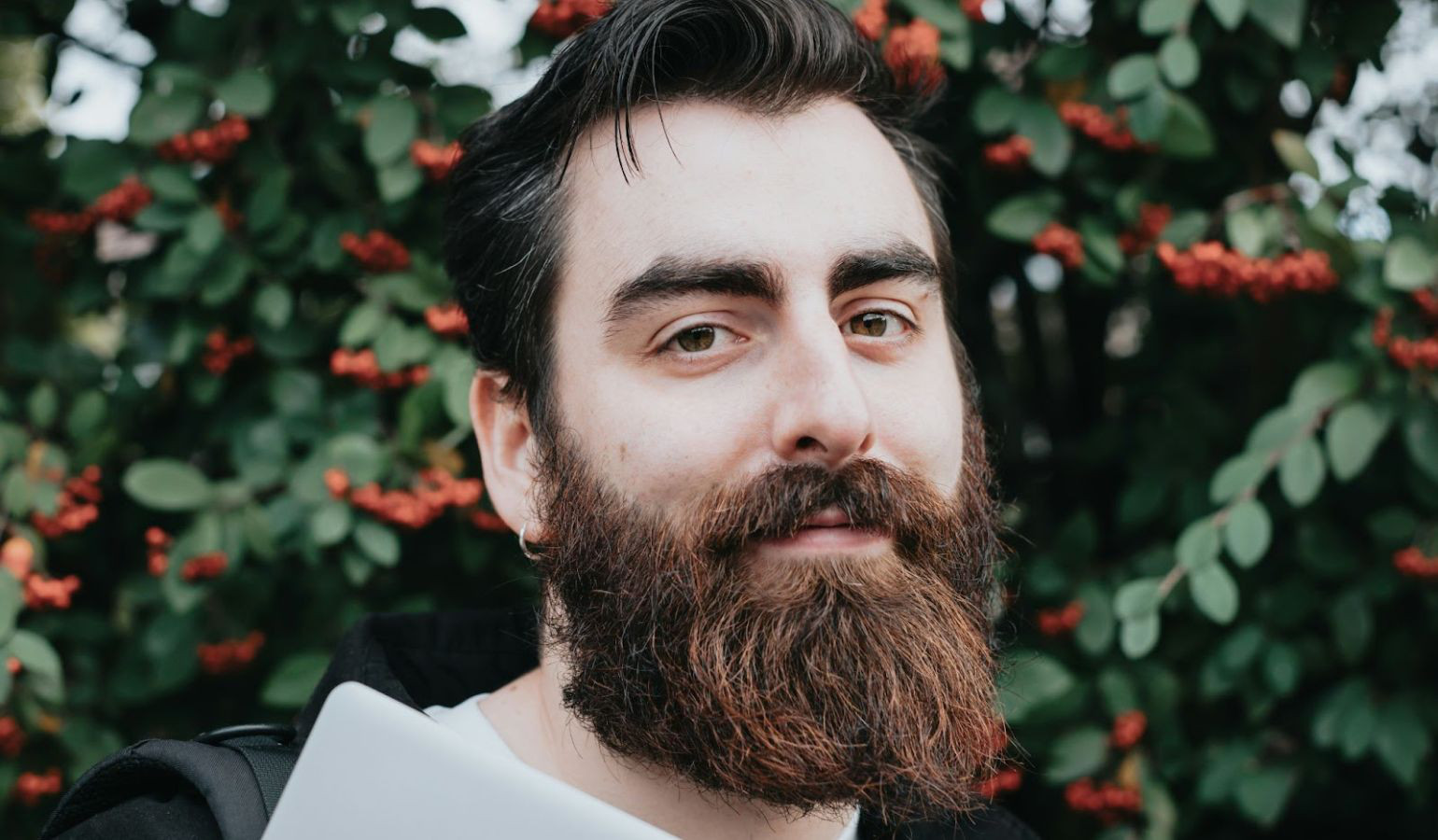 Young bearded man carrying a backpack and a laptop staring into the camera