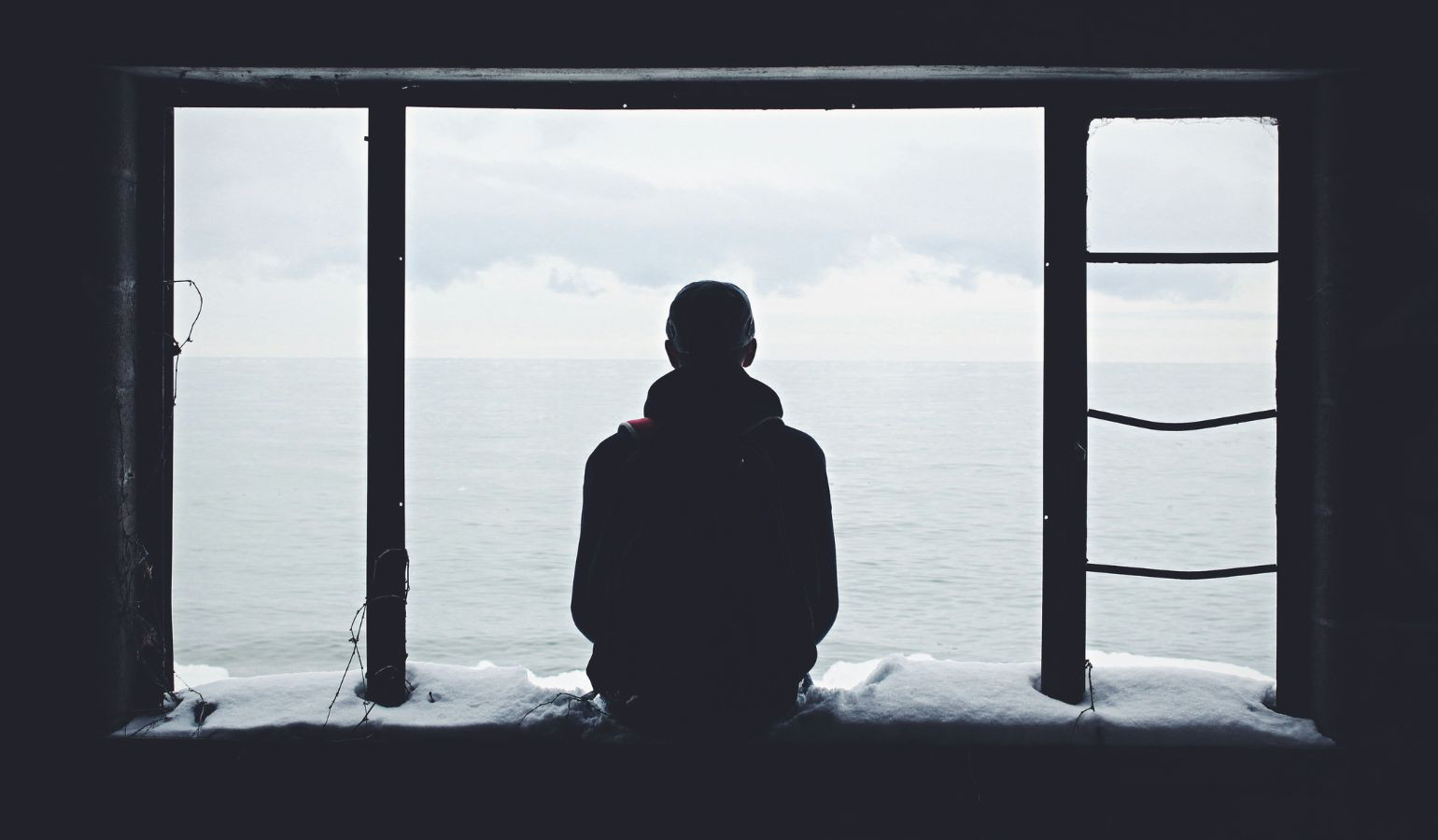 Silhouette of a young man sitting on a window looking out to the ocean on a cloudy day