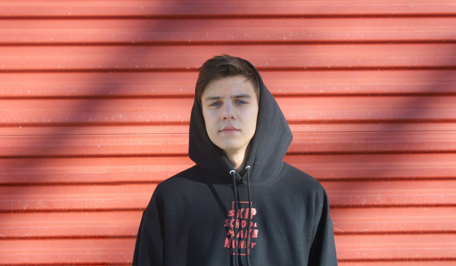 White teenager in a hoodie looking forward standing in front of a red wall