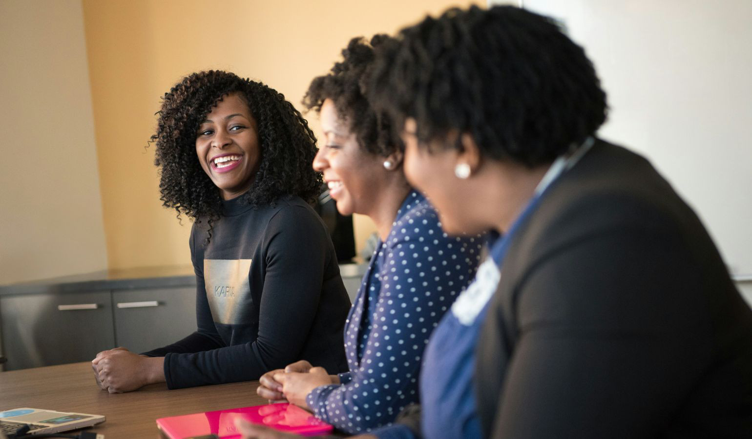 Three young African American women sitting at a business meeting smiling.