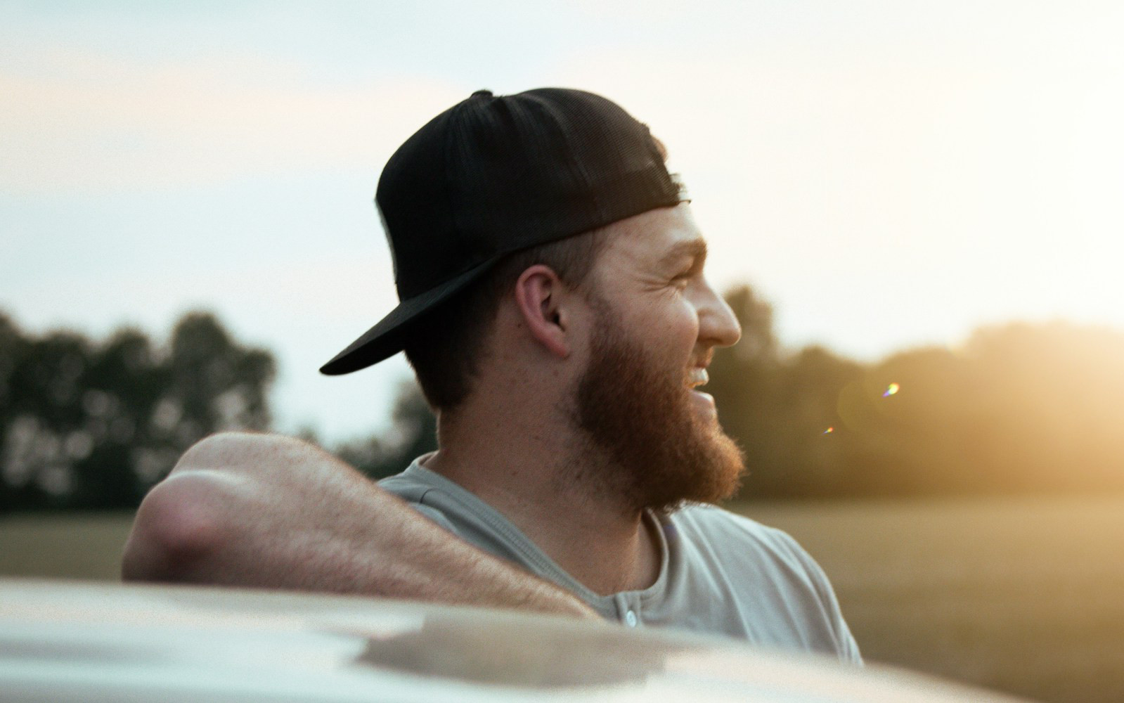 Young white male with ballcap Laughing and leaning against a car